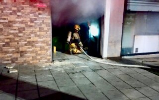 Un bombero se encuentra dentro de un local, controlando un incendio que se produjo en el interior de este, del local sale despedido humo por causa del agua que se le ha arrojado.